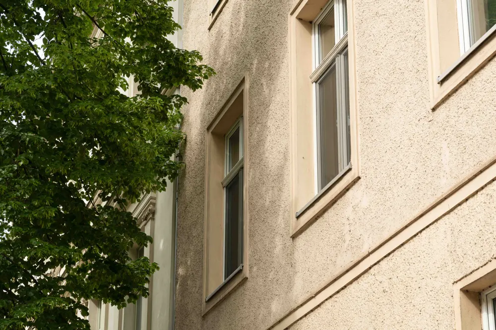 Close-up of a beige stucco building facade with tall rectangular windows next to a lush green tree.