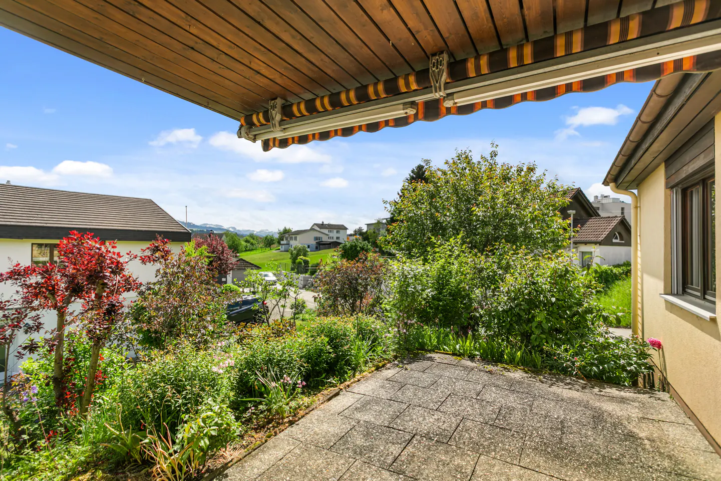 View from a stone patio with a striped awning, overlooking a lush garden and houses on a green hillside under a blue sky.