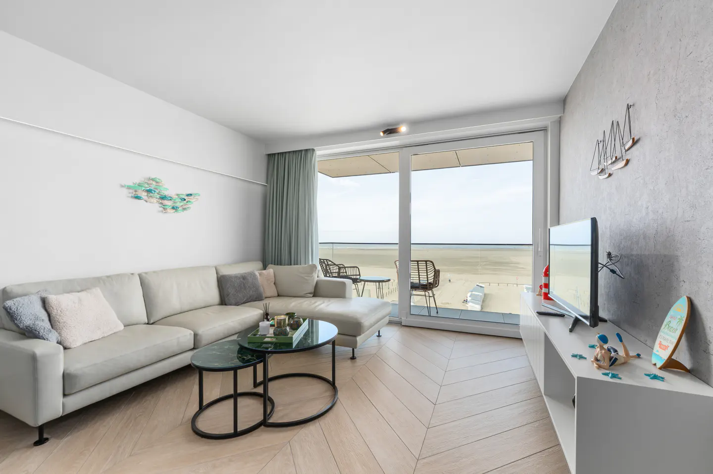 Bright living room with a beige sectional sofa, round coffee tables, and a view of the beach from a large sliding glass door.