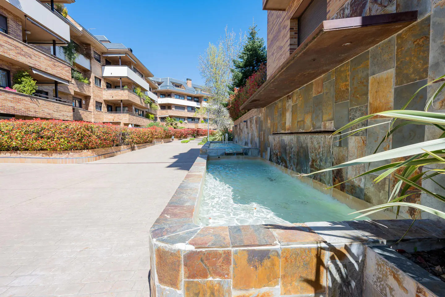 Exterior view of a stone fountain with water, surrounded by brick buildings and green plants on a sunny day.