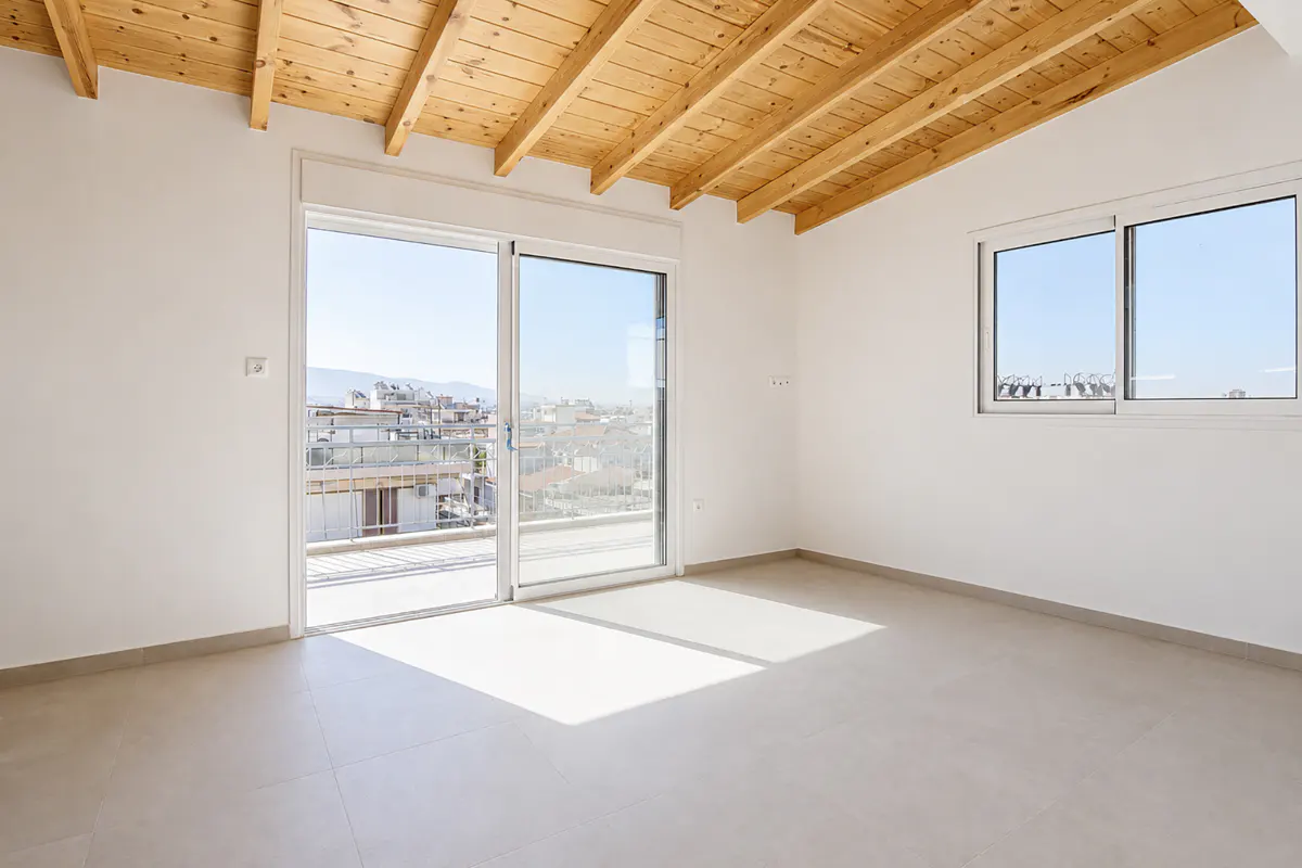 Empty room with white walls, light tile floors, a wooden beamed ceiling, and a balcony door overlooking a city view.