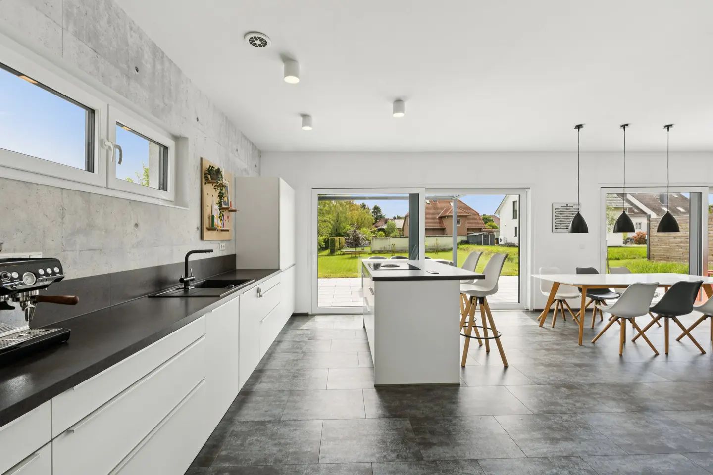 Bright, modern kitchen with white cabinets, black countertops, and gray tile flooring. Large windows overlook a green lawn and houses.