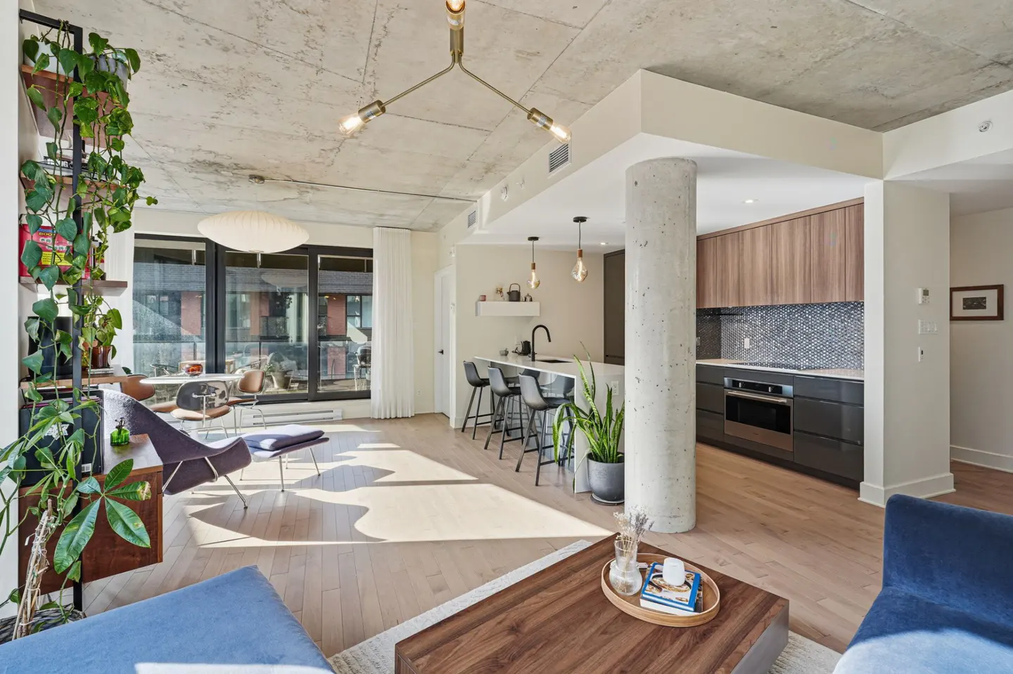 Open-concept living space with concrete ceiling, wood floors, and modern furniture. Kitchen with island and bar stools visible.