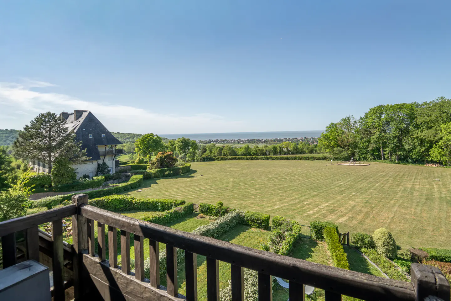 View from a dark wood balcony overlooking a large green lawn, trees, and a house with a dark roof. The ocean is in the distance under a blue sky.