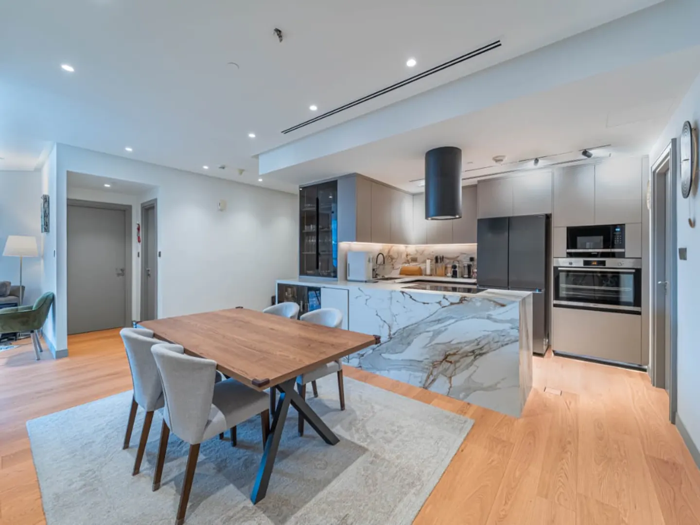 Modern kitchen with marble island, wooden dining table, grey chairs, and light wood floors in an open-plan apartment.