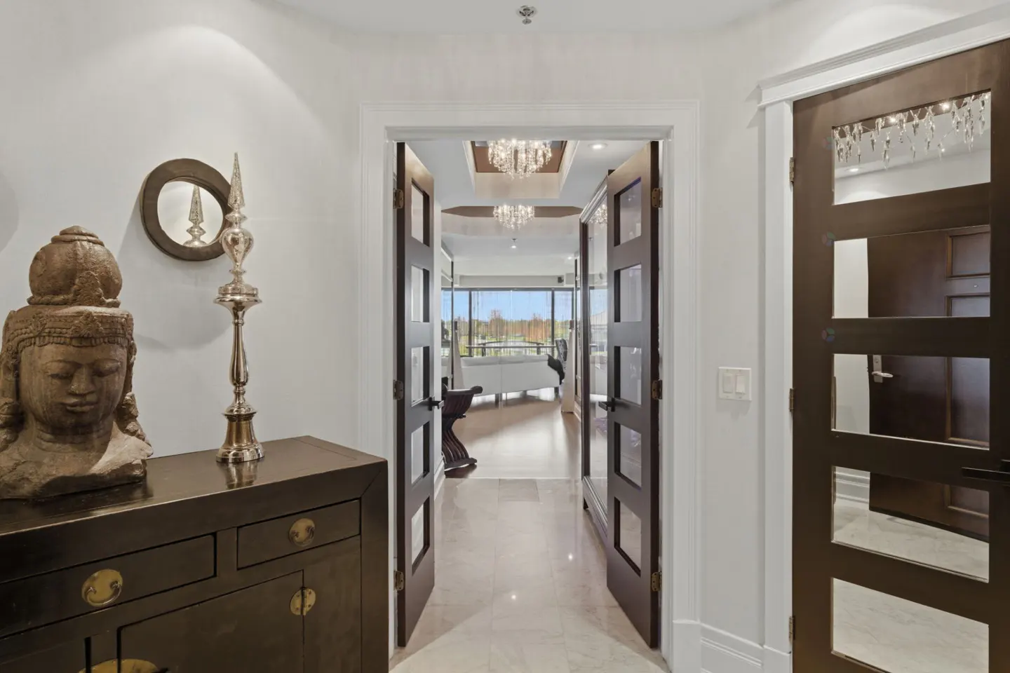 Entryway with a dark wood cabinet, Buddha statue, and silver candlestick. Double doors lead to a living room with a chandelier and a view of trees.