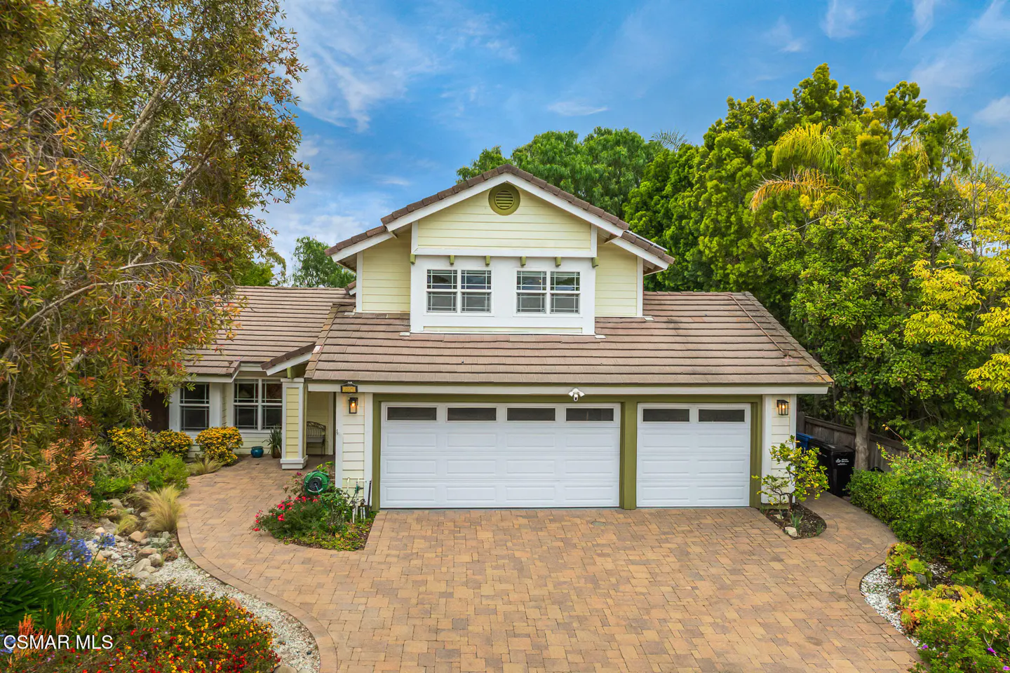 Two-story house with a brown tile roof, pale yellow siding, and a two-car garage. A brick driveway and walkway lead to the house.