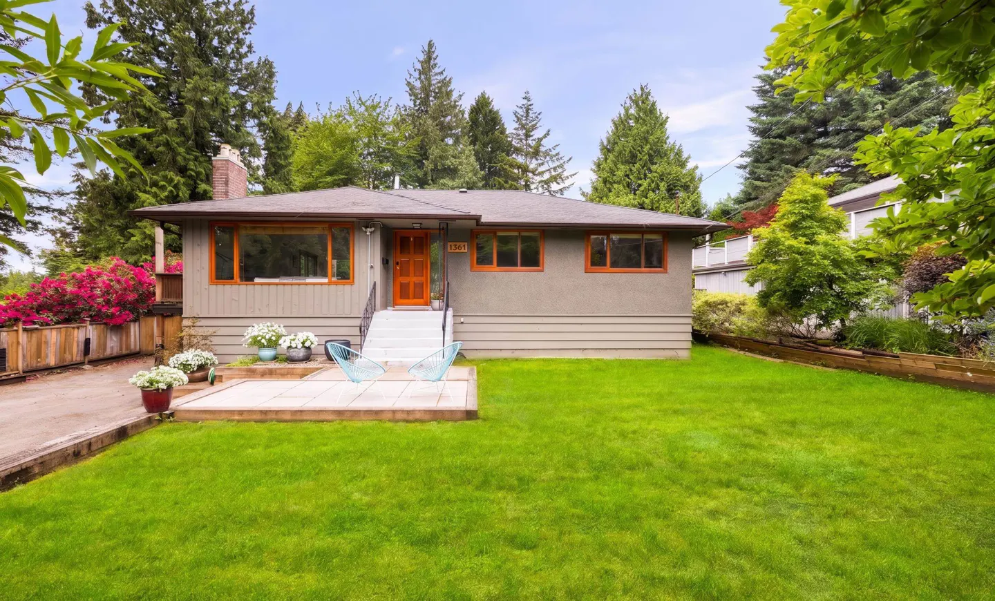 Single-story grey house with orange trim, a front patio with two blue chairs, and a large green lawn under tall trees.