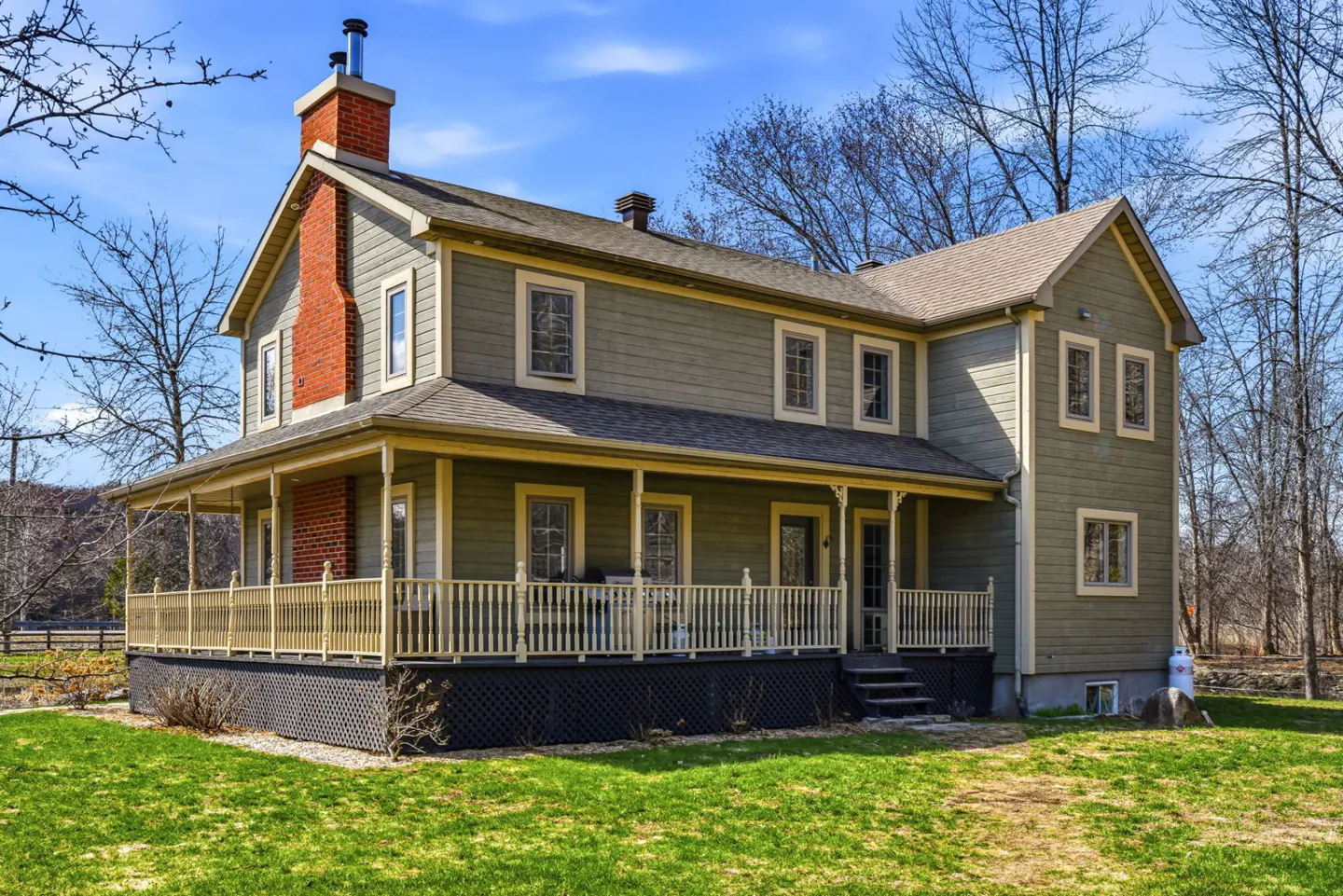Two-story olive green house with a brick chimney and a porch, set on a green lawn under a blue sky.