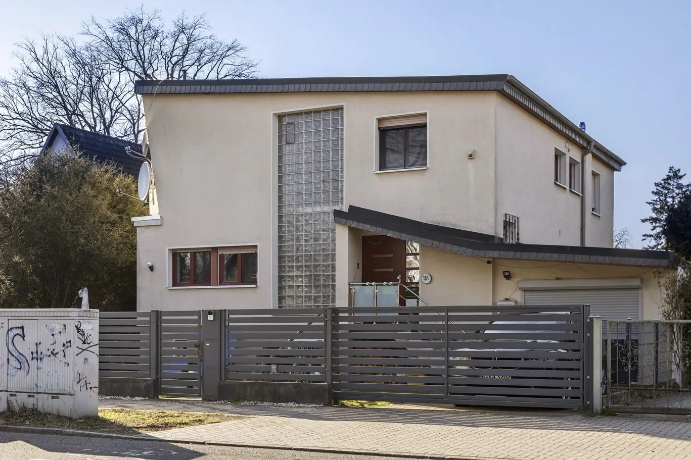 Two-story beige house with a gray horizontal slat fence and a garage. A glass block window is near the front door.