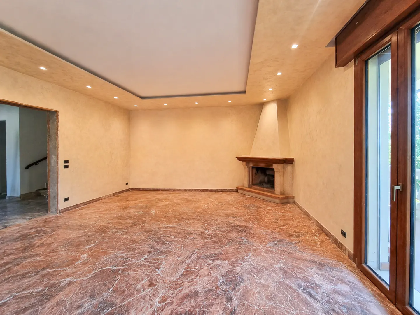 An empty living room with marble floors, beige walls, and a fireplace. A doorway and a glass door are visible.