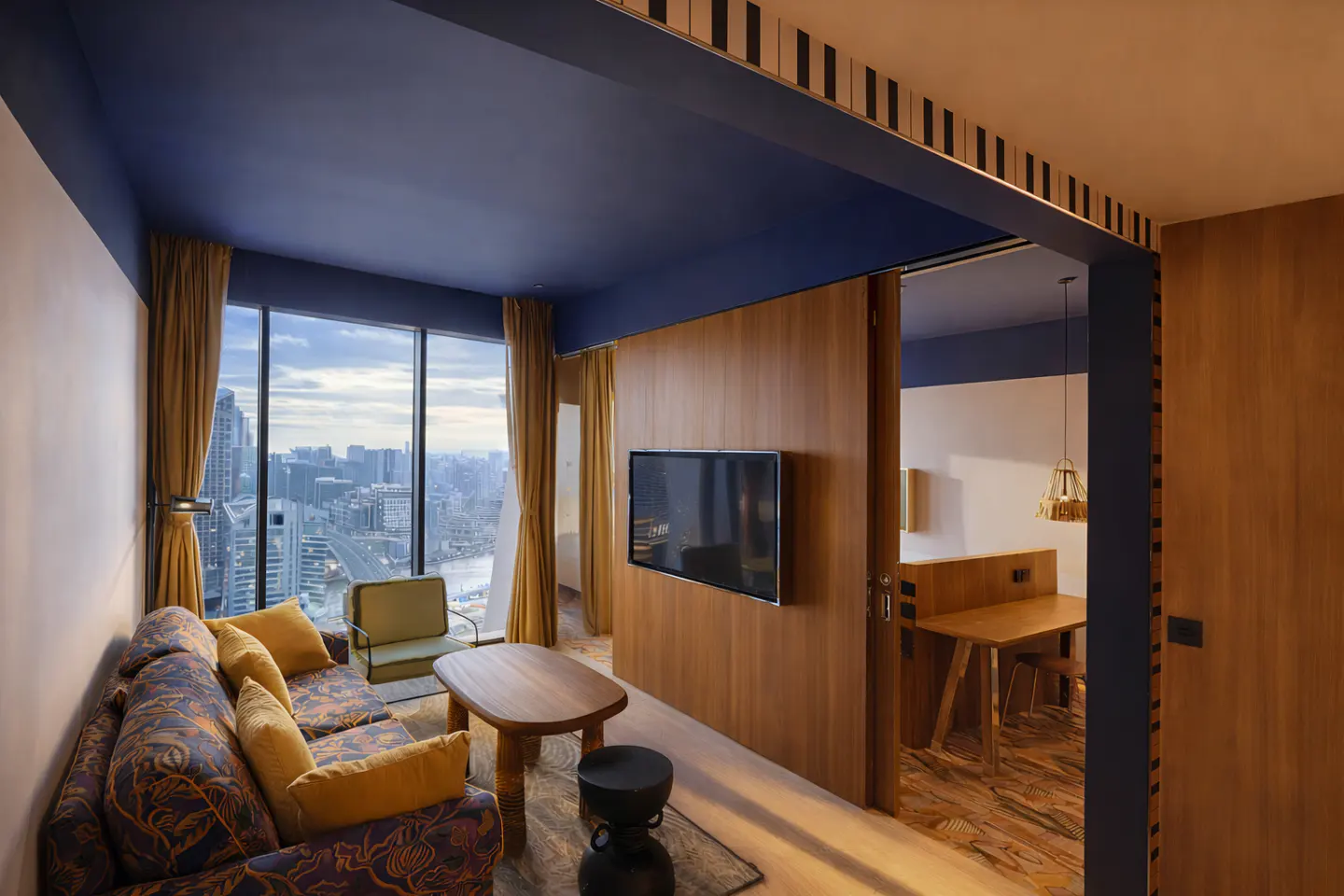 Hotel suite with city view. Sofa, chair, and table in the living area. A TV is mounted on a wood-paneled wall. A bedroom is visible through a doorway.