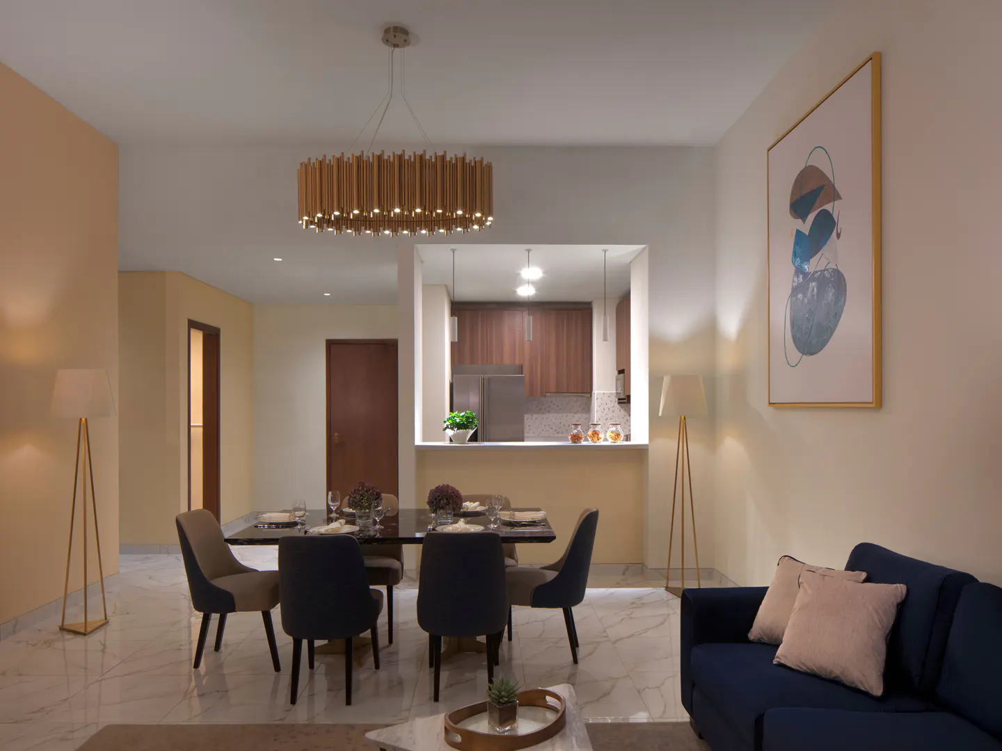 A modern apartment interior with a dining table, blue sofa, and open kitchen. A gold chandelier hangs above the table. Artwork adorns the walls.