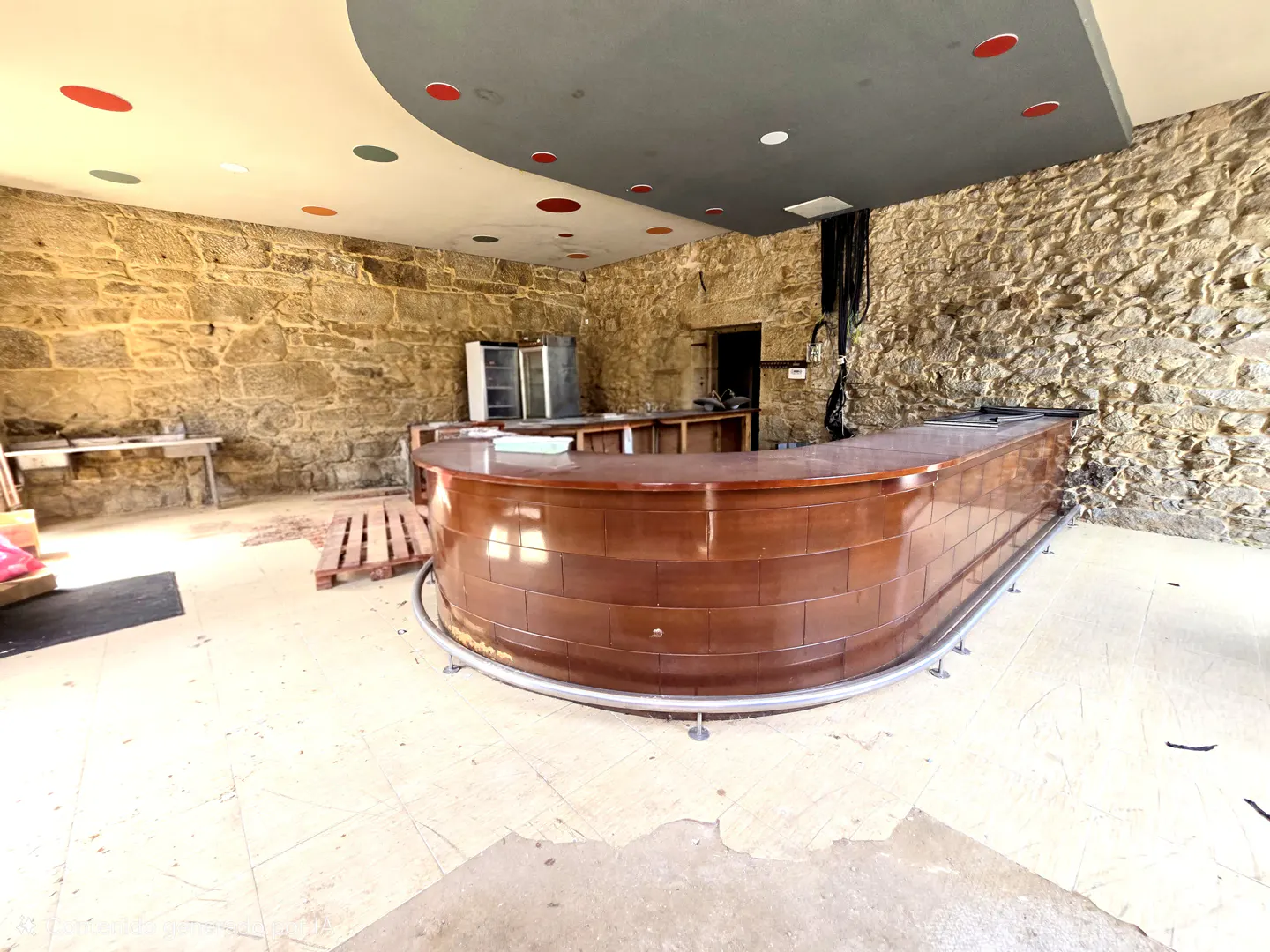 Interior view of a bar area with stone walls, a curved wooden bar, and a gray ceiling with red and green accents.