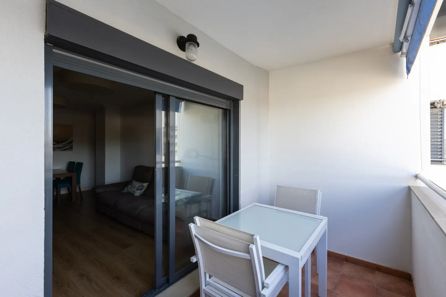Balcony with a white table and chairs, sliding glass doors to a living room with a brown sofa and dining table.