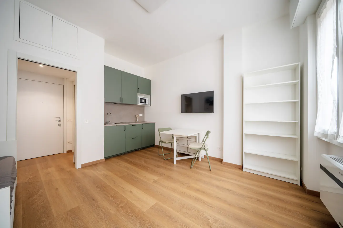 Bright studio apartment with wood floors, green kitchen cabinets, white table and chairs, TV, and white bookshelf.