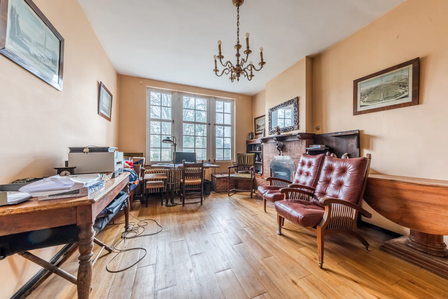 A home office with wood floors, a fireplace, and two red leather chairs. A desk sits in front of a large window.