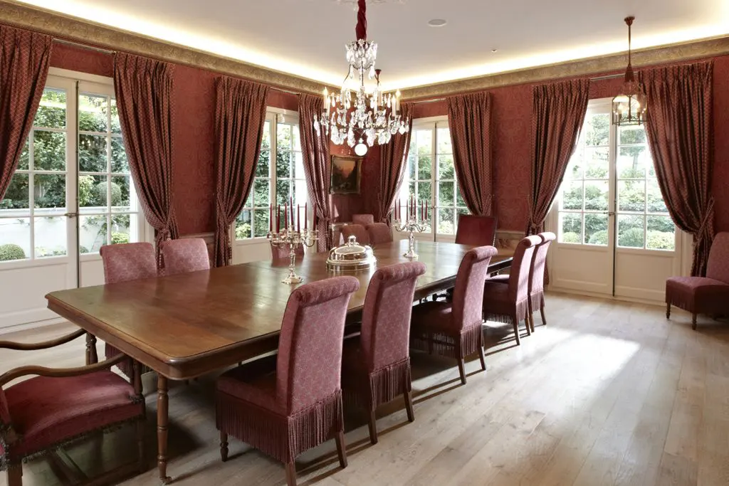 Formal dining room with a long wooden table, red patterned walls and curtains, and a crystal chandelier. Windows overlook a green garden.