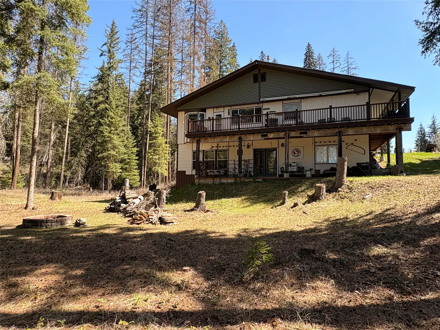 Two-story house with a deck, surrounded by tall trees and a grassy yard. A fire pit and wood pile are in the foreground.