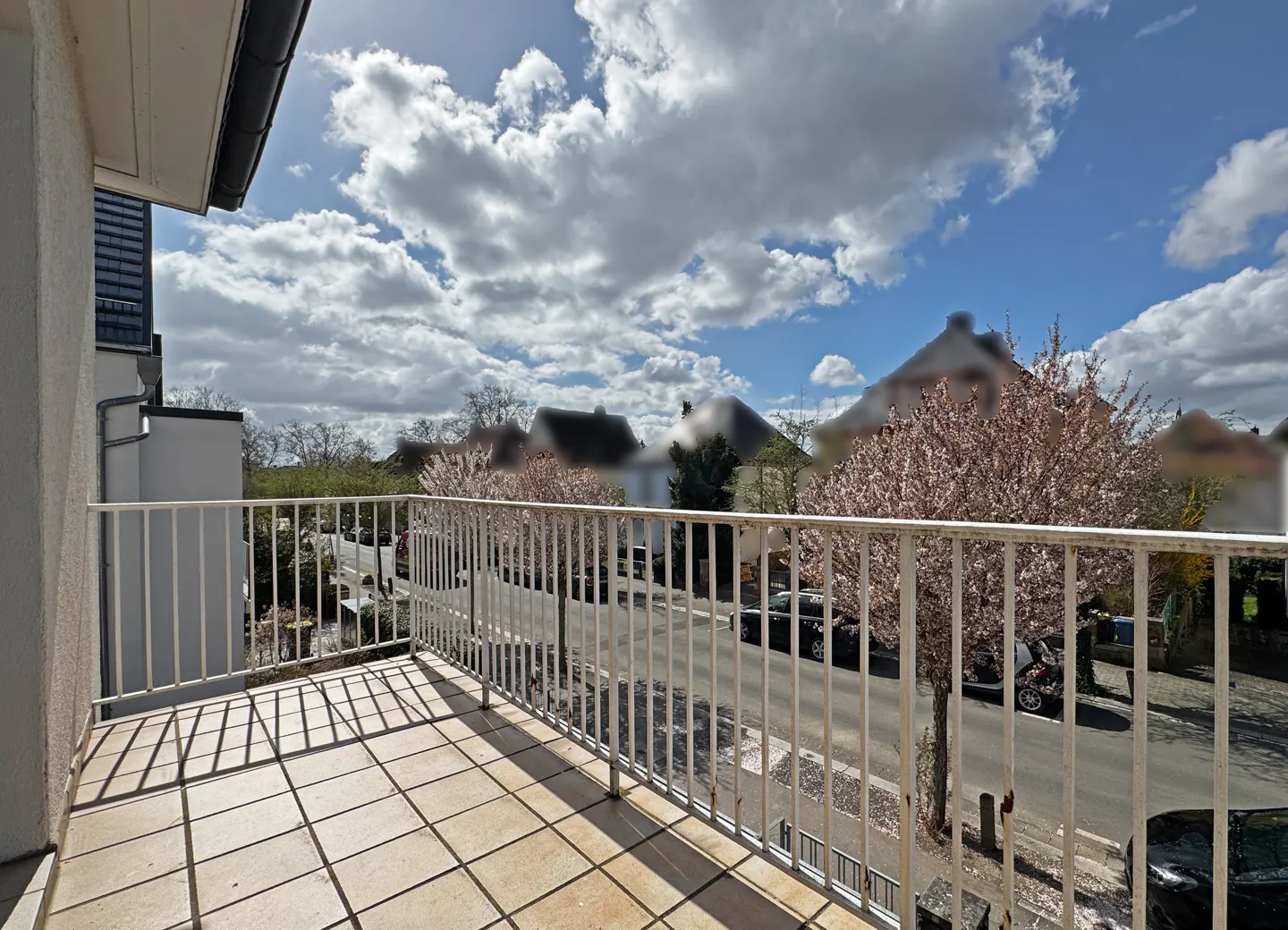 Balcony view with white railings, tan tiled floor, and street view. Flowering trees line the street under a cloudy blue sky.