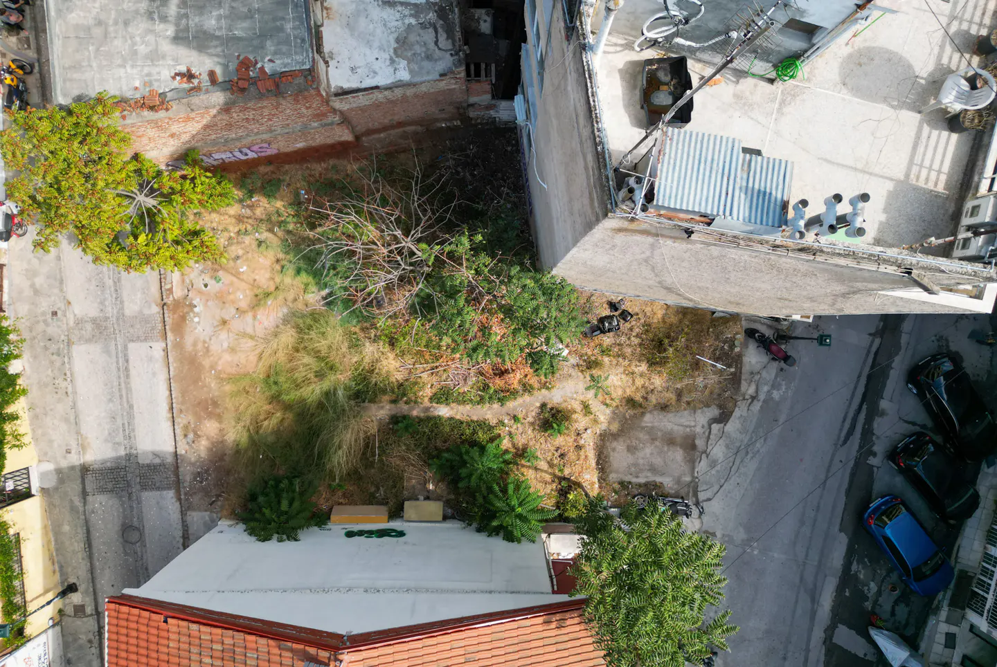Aerial view of a vacant lot surrounded by buildings and streets with parked cars. The lot has overgrown grass and a few trees.