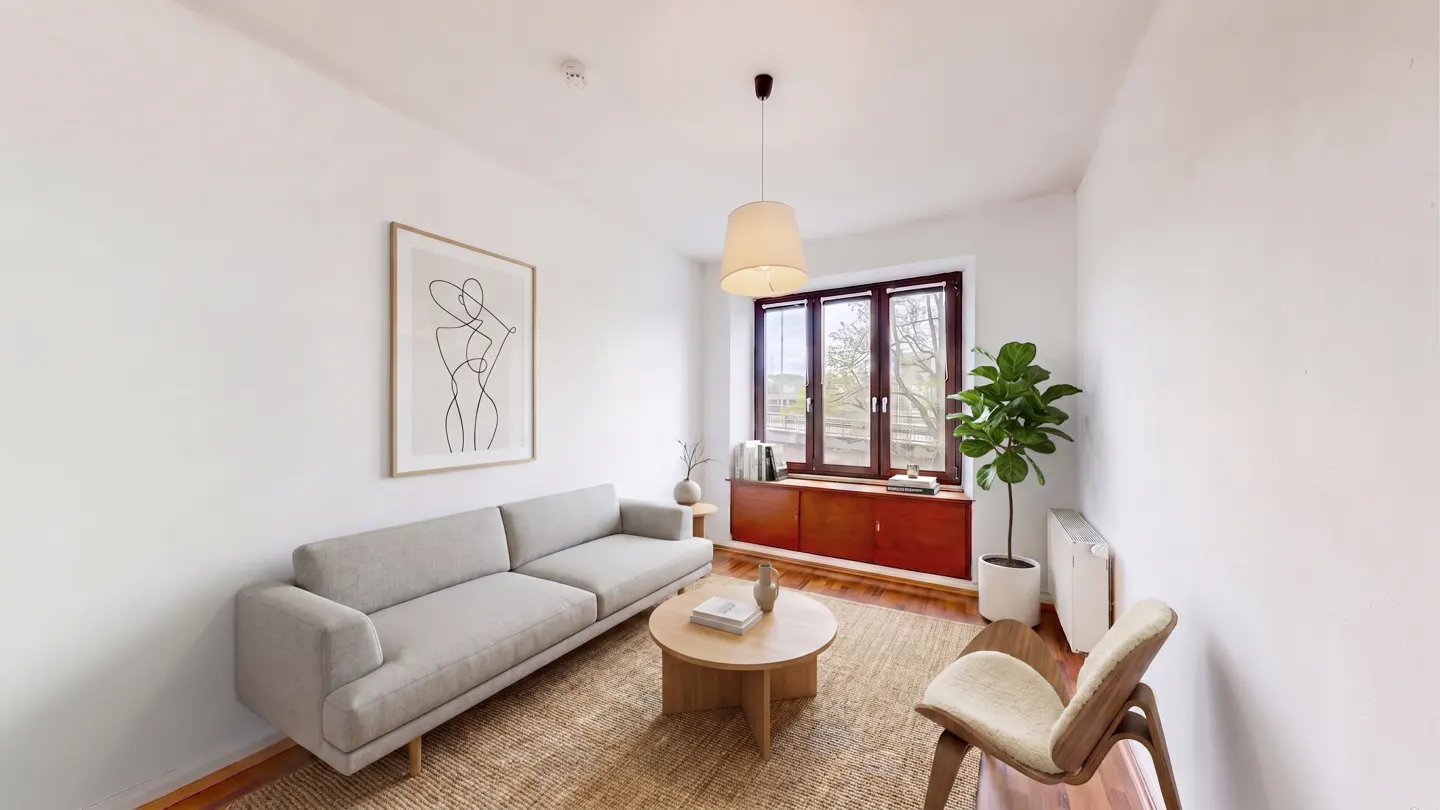 Bright living room with a gray sofa, round wood table, and a tan rug. A modern art print hangs on the white wall. A window with a red cabinet sits in the background.