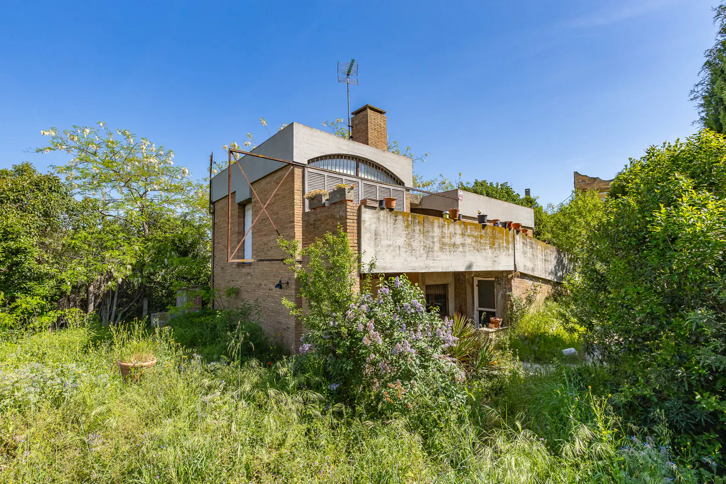 Exterior view of a two-story brick house with a concrete balcony, surrounded by overgrown greenery under a clear blue sky.