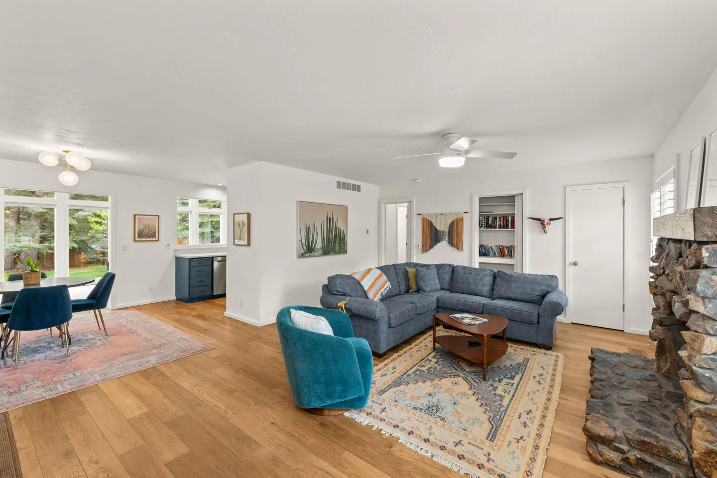 Open-concept living room with wood floors, blue sectional sofa, stone fireplace, and dining area with large windows.