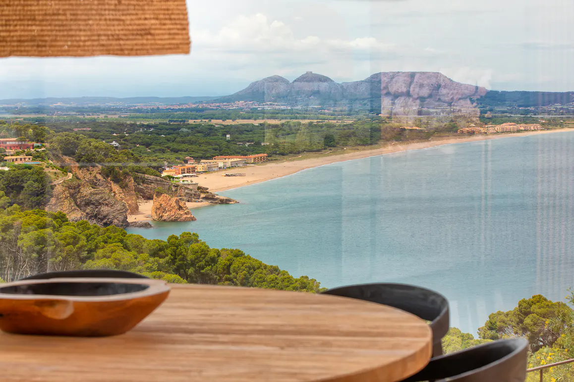 View from a dining room overlooking a sandy beach, turquoise sea, and distant mountains under a cloudy sky.