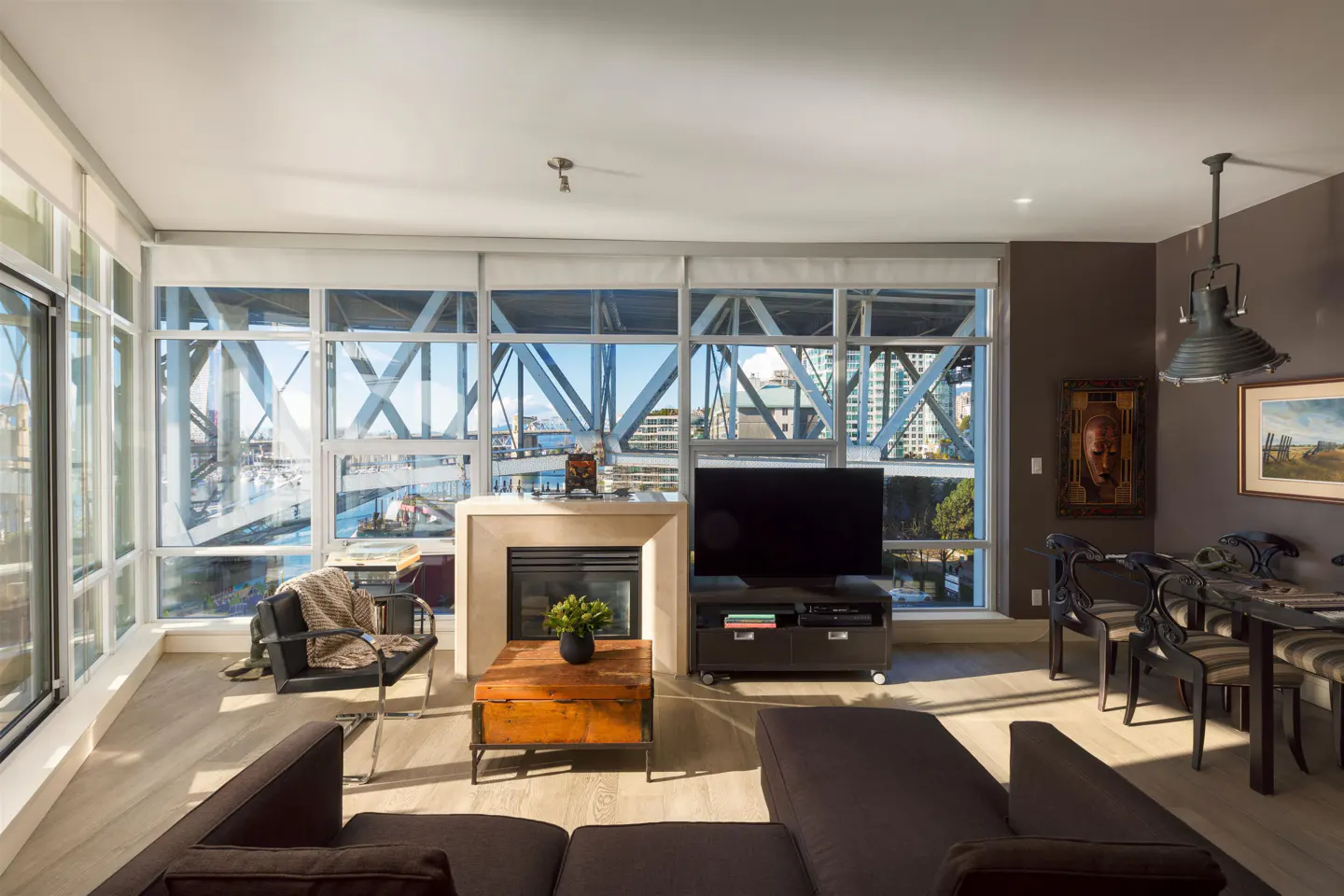 Living room with floor-to-ceiling windows showing a bridge and city view. A brown sofa, fireplace, and dining table are visible.