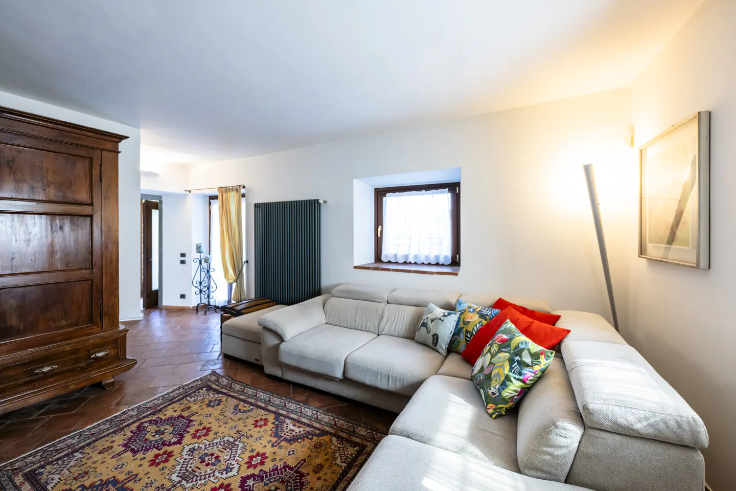 Living room with a beige sectional sofa, colorful pillows, and a patterned rug on terracotta tile floor. A dark wood armoire stands near an open doorway.
