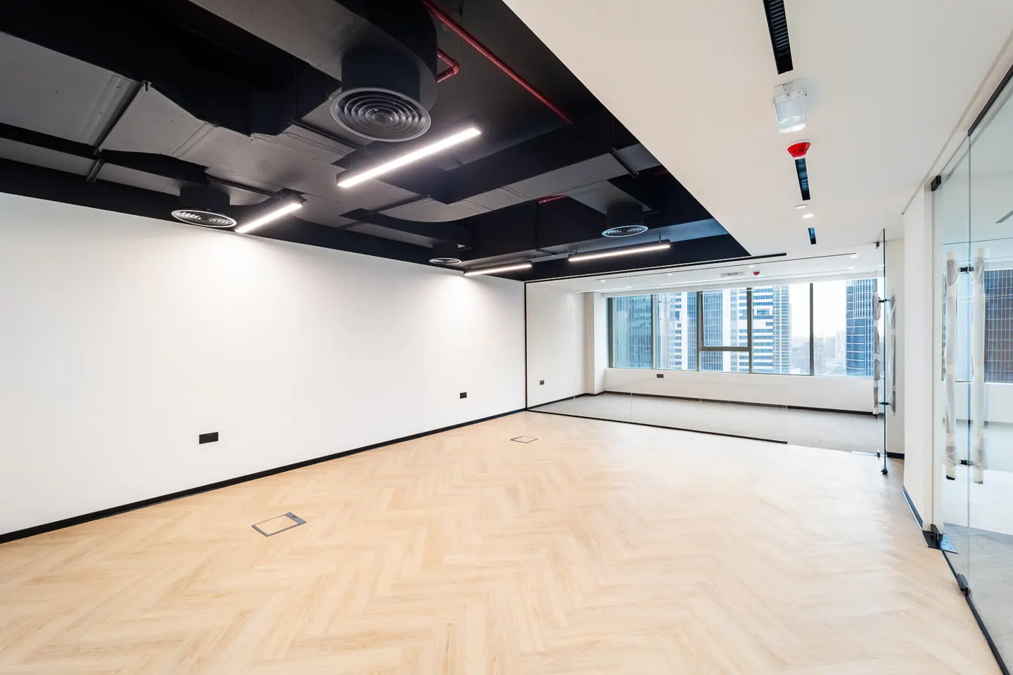 An empty office space with herringbone wood floors, white walls, and a black ceiling with exposed ductwork and linear lighting. A window shows a city skyline.
