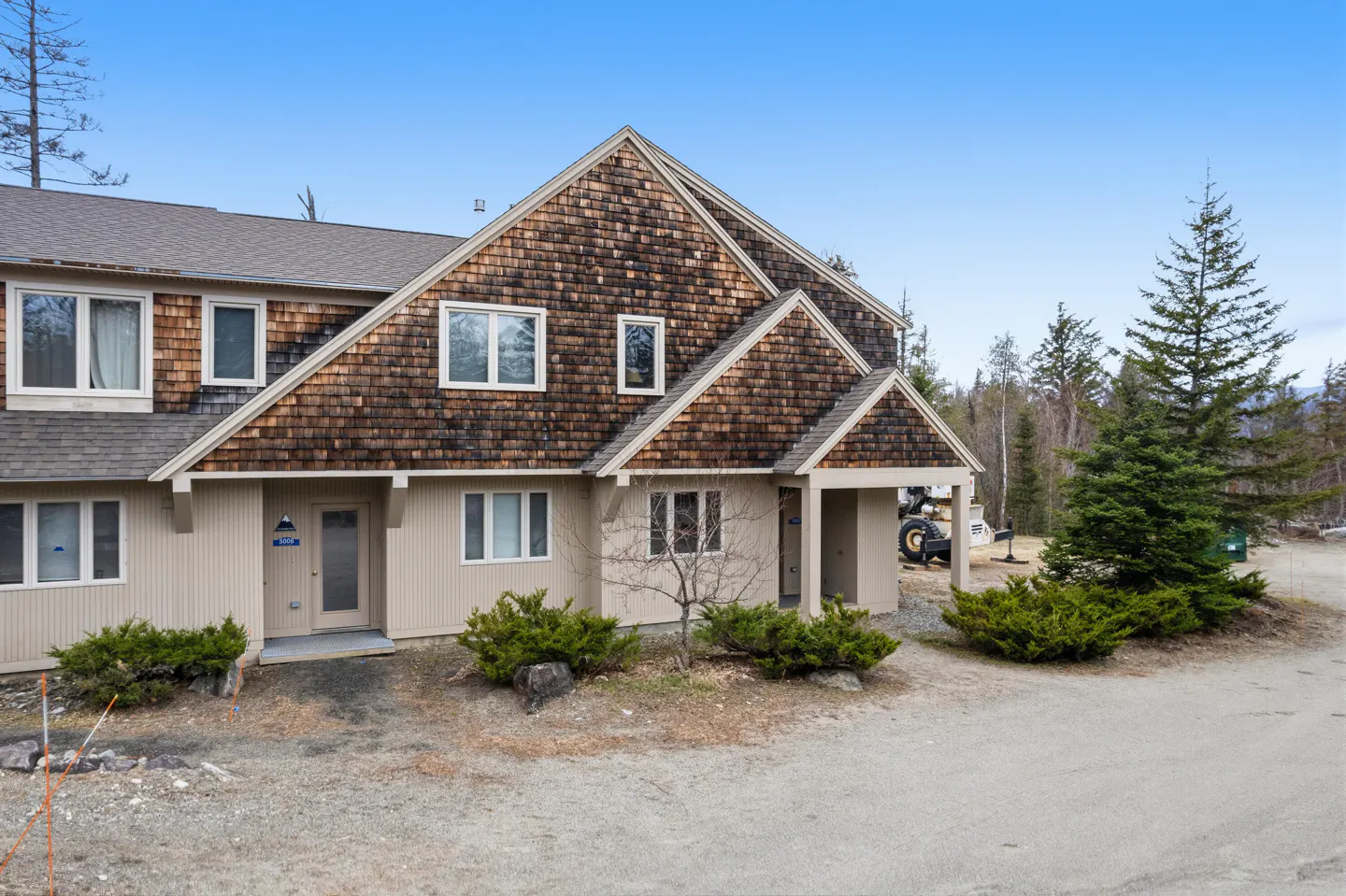 Exterior view of a two-story townhouse with brown shingle siding and a beige facade under a blue sky. Shrubs line the front.