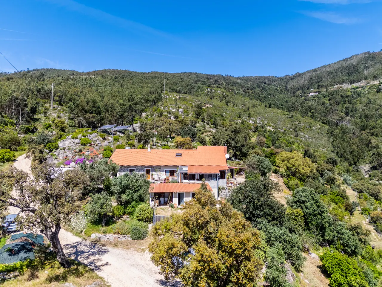 Exterior view of a stone house with an orange tile roof, surrounded by trees and a hillside under a blue sky.