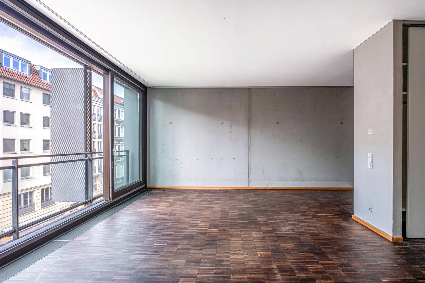 Empty room with dark parquet floor, gray walls, and large windows with a balcony overlooking a city building.