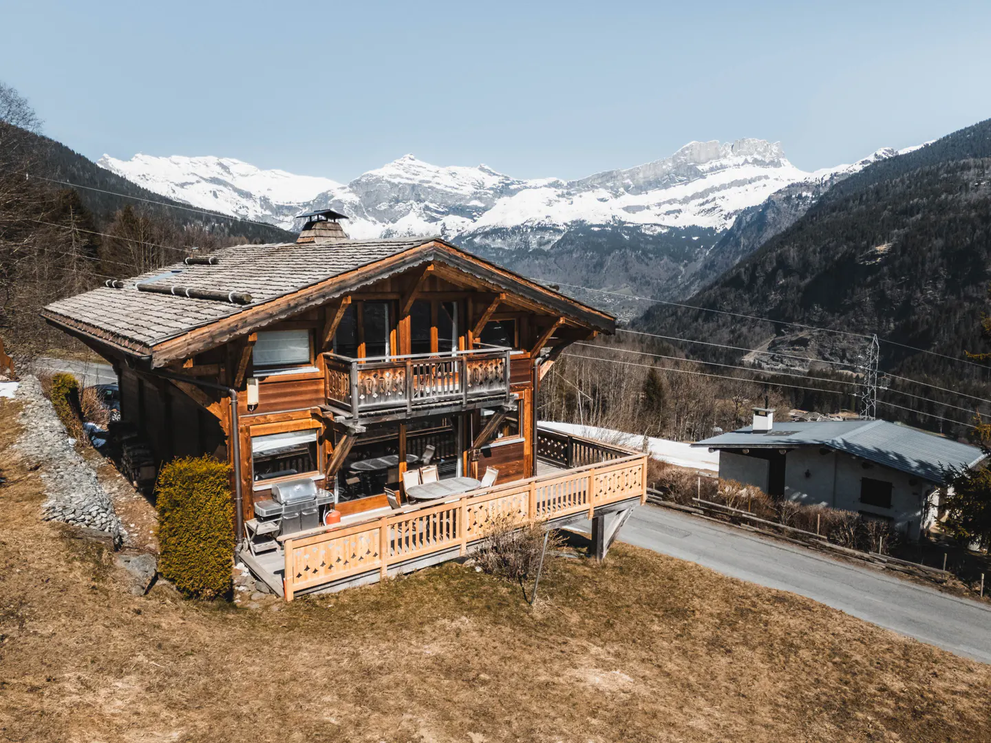 A wooden chalet with a balcony overlooks snow-capped mountains under a blue sky.