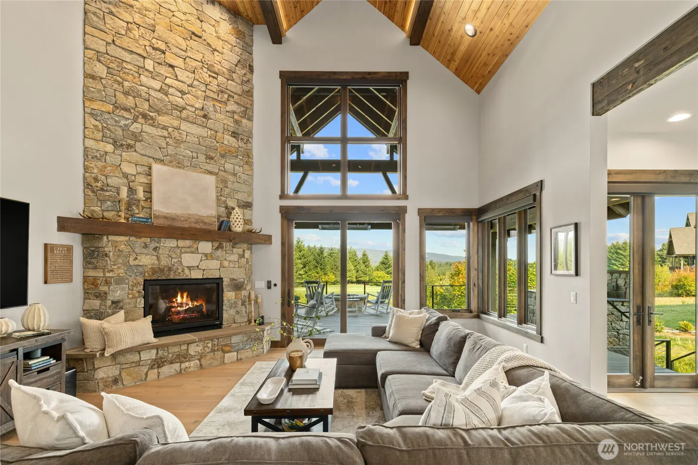 Living room with stone fireplace, wood beams, and large windows overlooking a green landscape. A gray sectional sofa sits in the foreground.