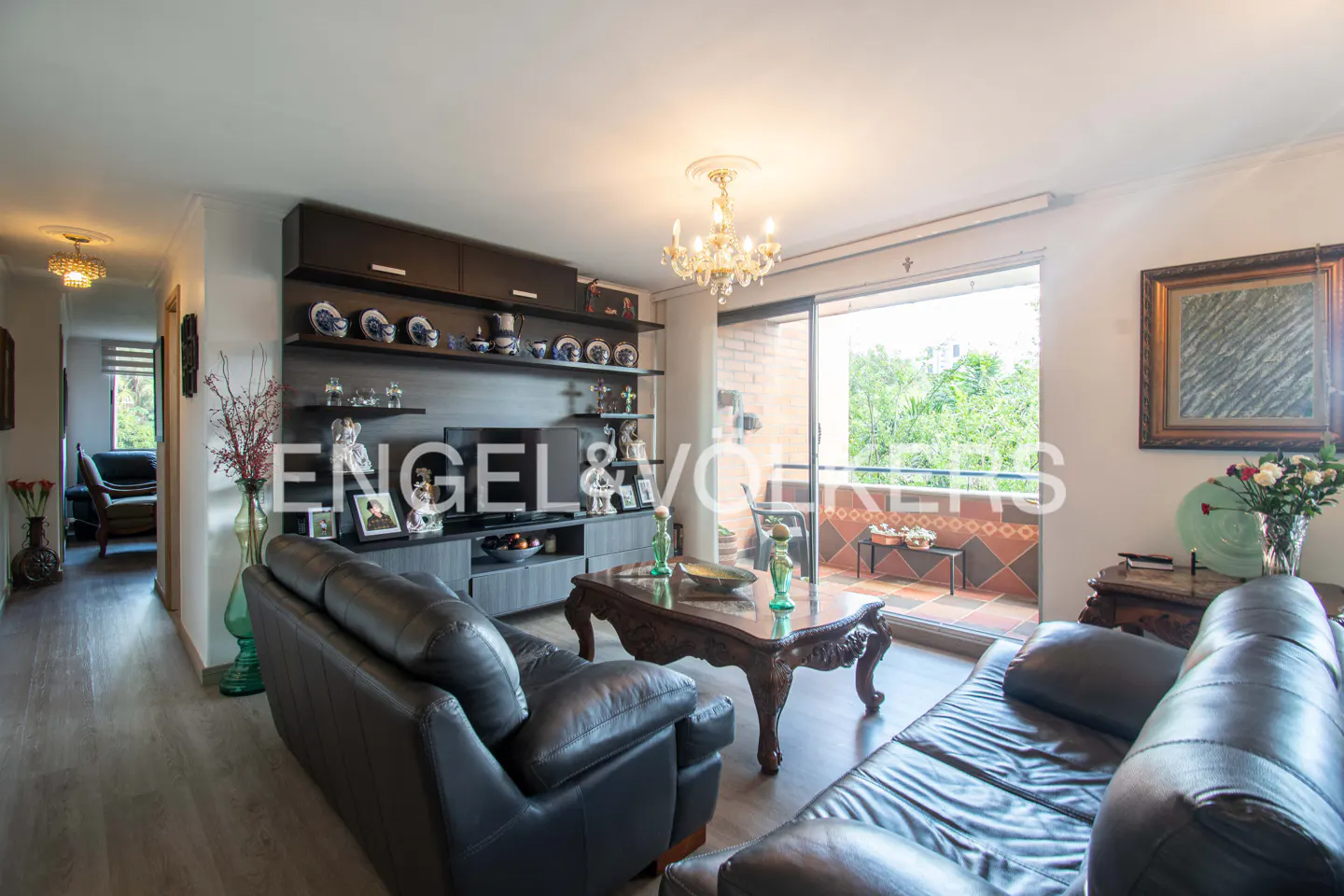 Living room with black leather sofas, dark wood shelves with decorative plates, and a balcony with green trees in the background.