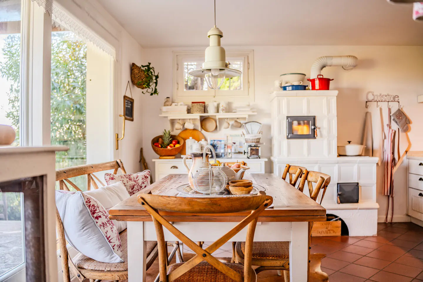 Bright kitchen with a wooden table, chairs, and a white tiled stove with a fire burning inside. Fruit bowl and dishes are on the table.