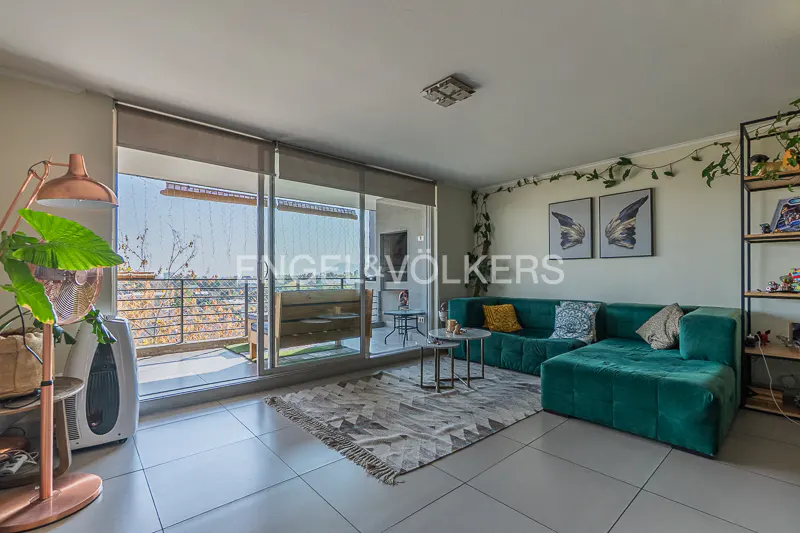 Living room with a green velvet sofa, patterned rug, and balcony with a view. Copper lamp and shelving unit add warmth.