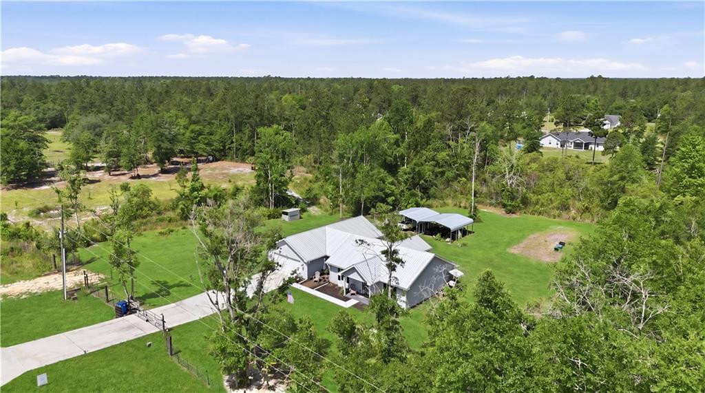 Aerial view of a gray house with a metal roof, surrounded by green trees and grass under a blue sky.