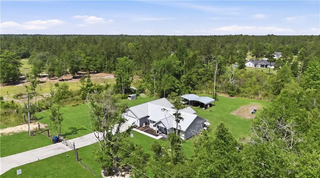 Aerial view of a gray house with a metal roof, surrounded by green trees and grass under a blue sky.
