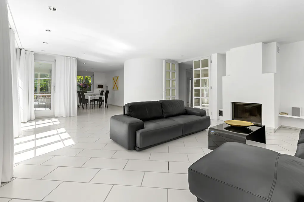 Bright, modern living room with white tile floors, black leather sofas, a fireplace, and a dining area in the background.