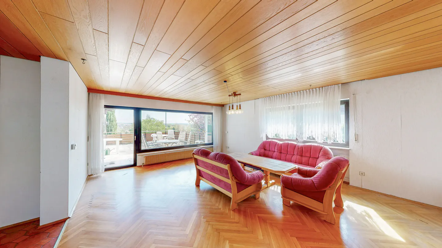 A bright living room with wood floors, a wood ceiling, and red sofas. A large window shows a patio with outdoor furniture.