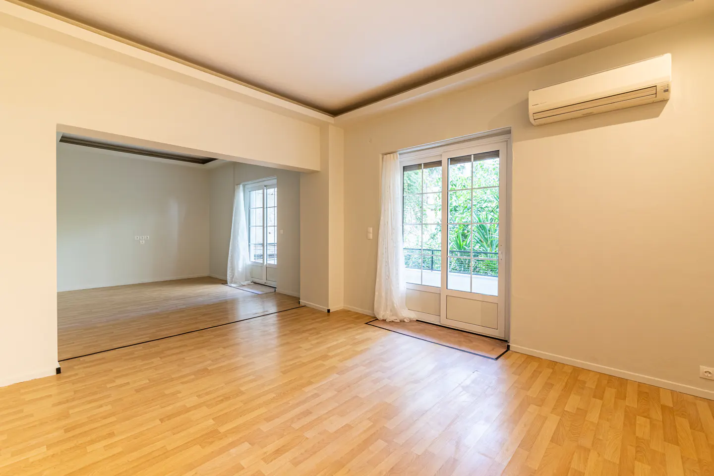 Bright, empty room with wood floors, cream walls, and white trim. Sliding glass doors lead to a green garden. An air conditioner is mounted high on the wall.