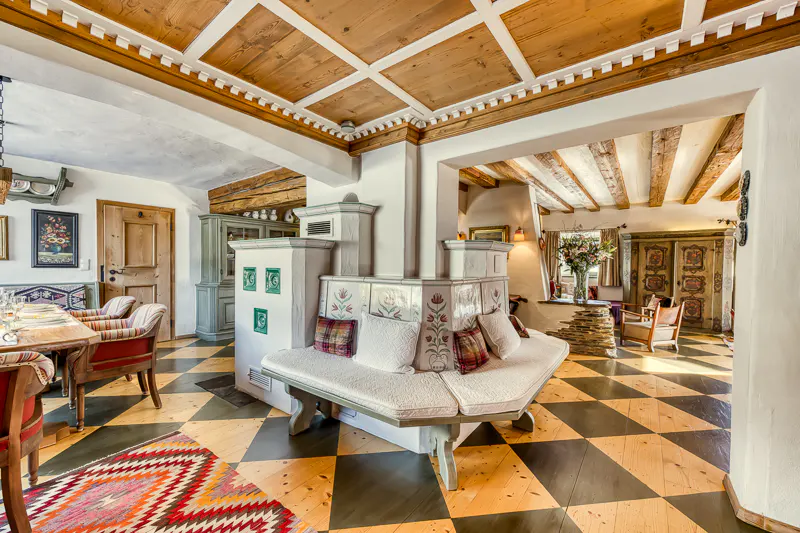 A cozy living room with a tiled stove bench, wooden ceiling, and checkered floor.
