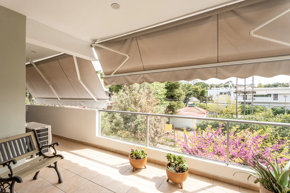 A sunny balcony with beige awnings, a bench, potted plants, and a glass railing overlooking trees and buildings.