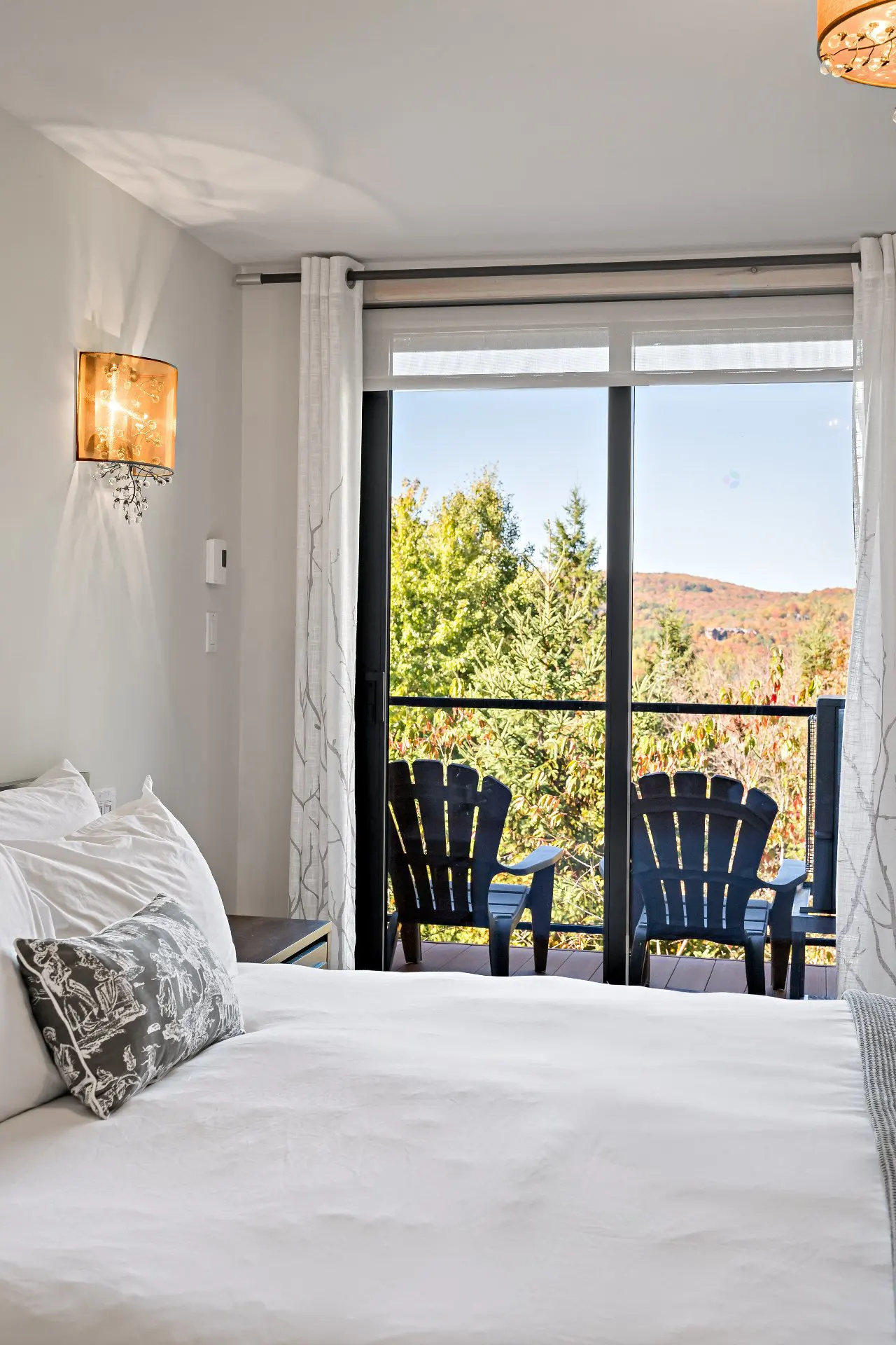 Bedroom with white linens, decorative pillow, and balcony with two black Adirondack chairs overlooking trees.
