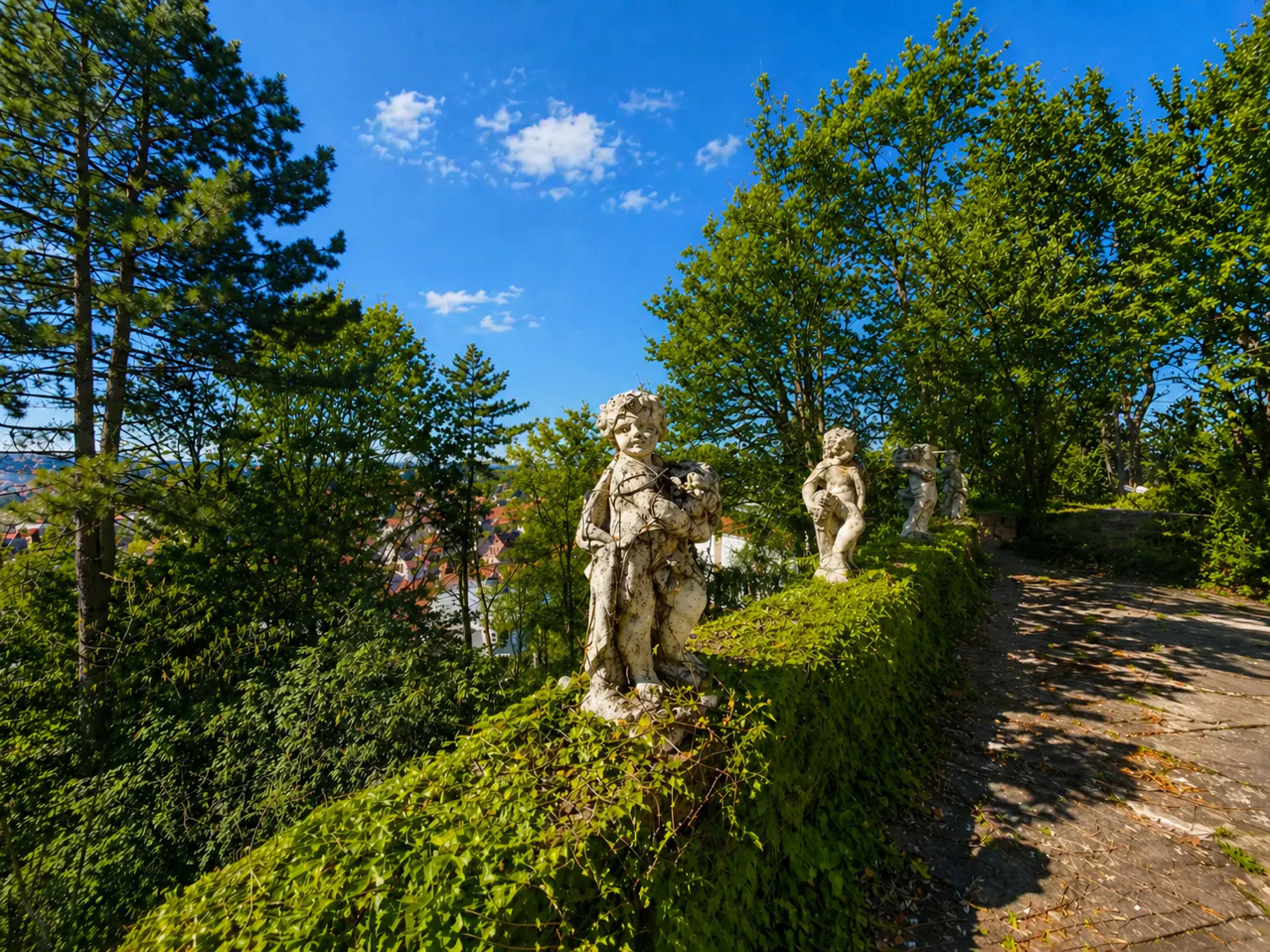Outdoor shot of cherub statues lining a green, ivy-covered wall, with trees and a blue sky in the background.