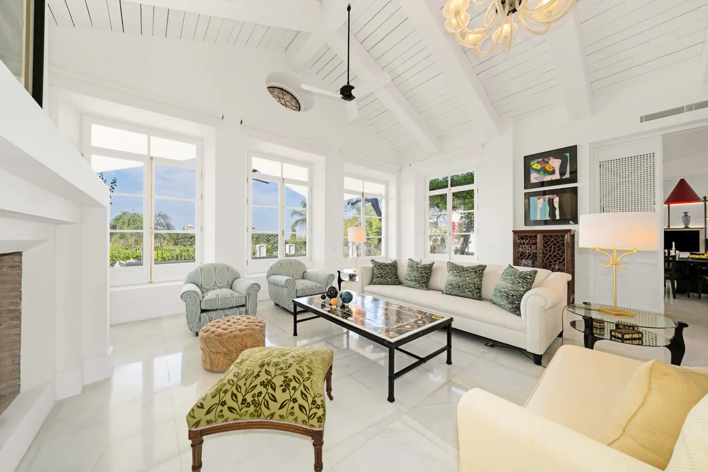 Bright living room with white walls, marble floors, and high ceilings. Large windows offer a view of greenery. Furnished with sofas, chairs, and a decorative coffee table.